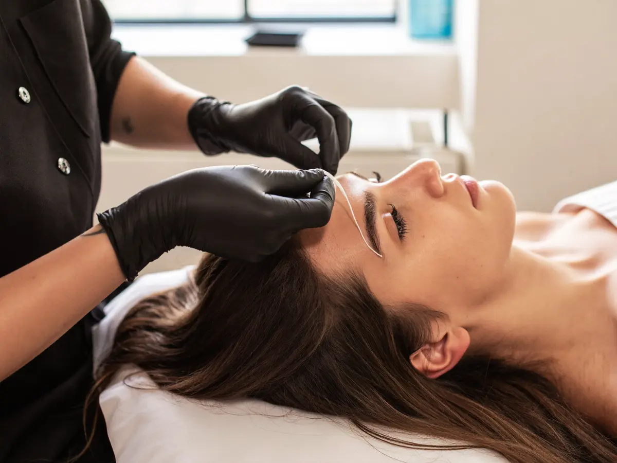 Esteticista depilando las cejas con hilo a una clienta tumbada en la camilla